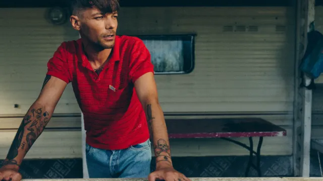 Louis Tomlinson de camisola vermelha numa varanda branca em frente a uma autocaravana.